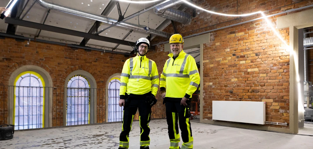 Interiör av en byggarbetsplats med tegelväggar, takinstallationer och två personer i skyddskläder.